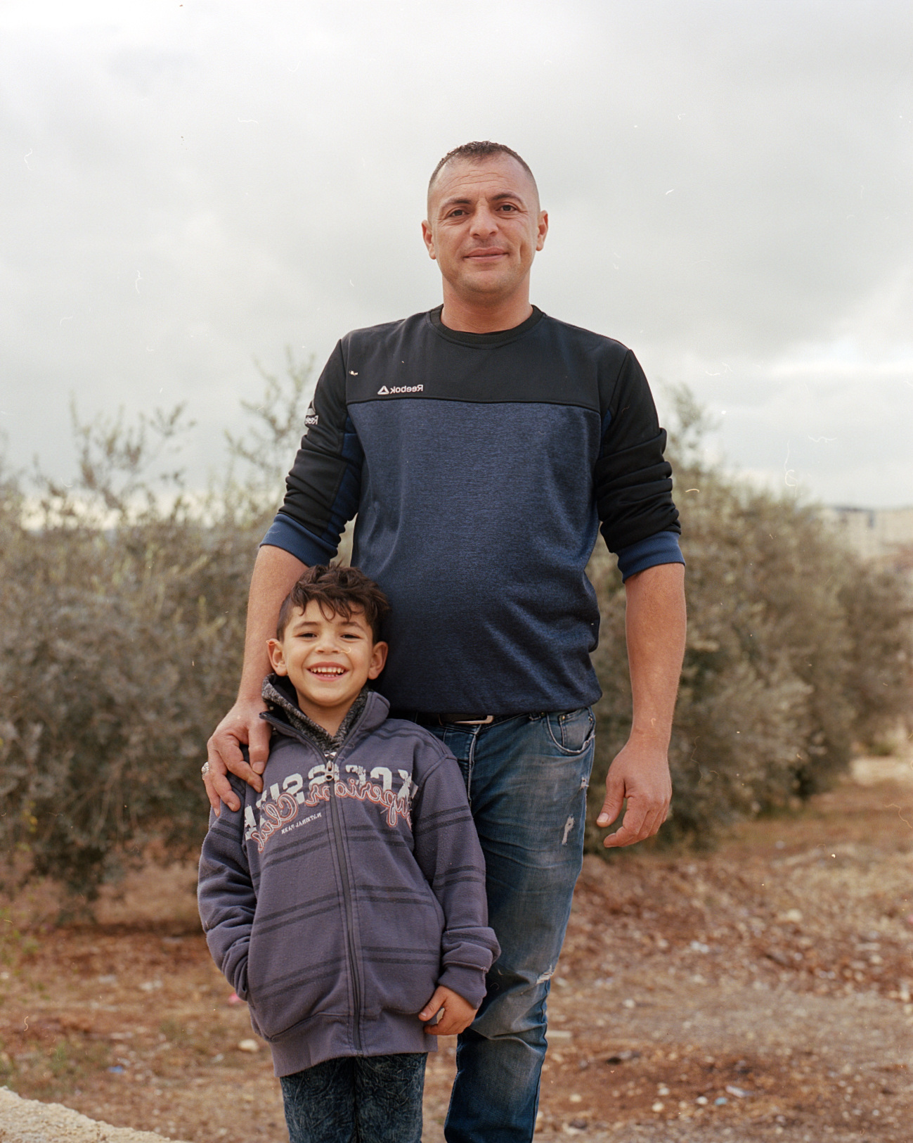 An adult and child standing together beside olive trees.