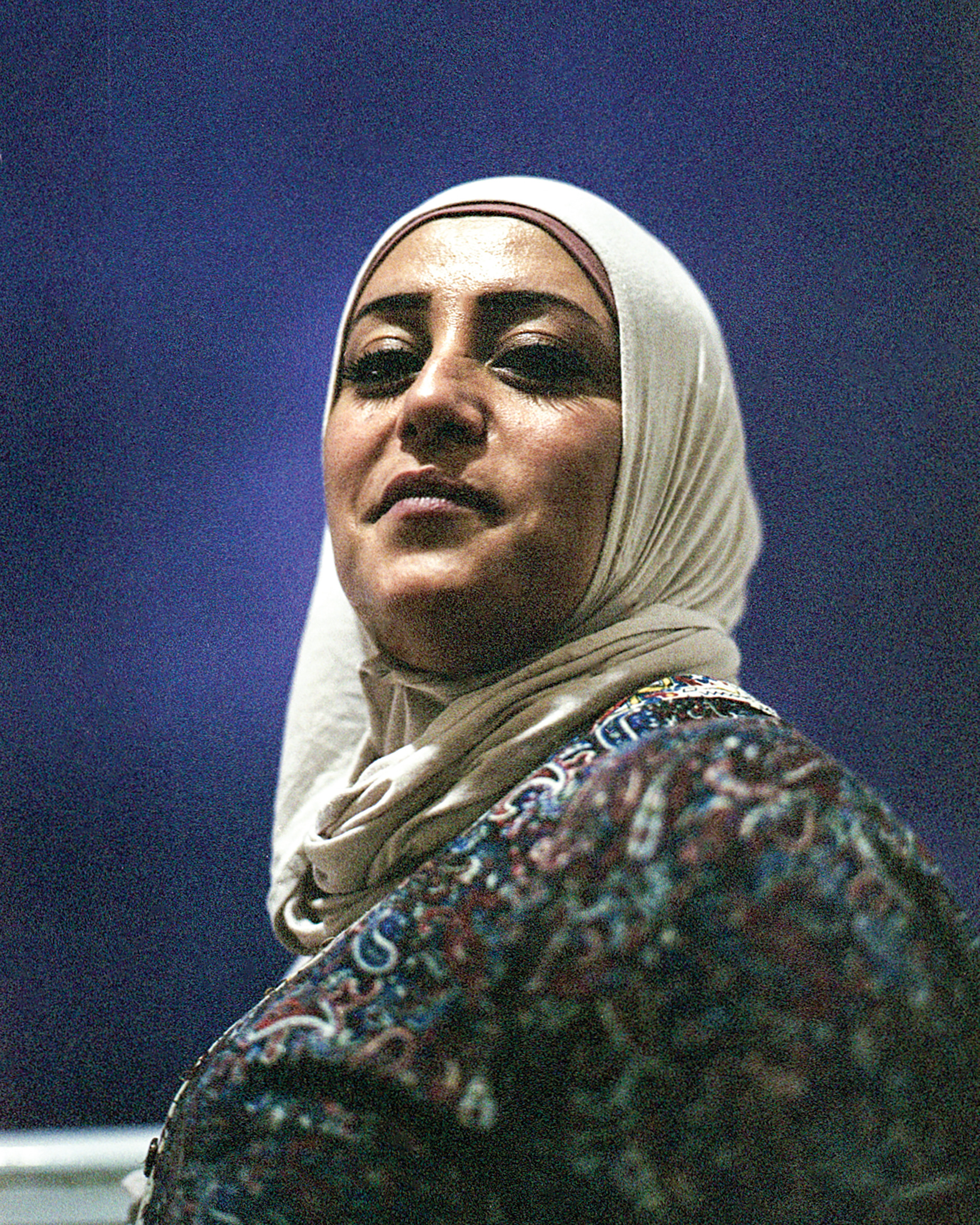 A portrait of a woman wearing a light headscarf against a blue wall.