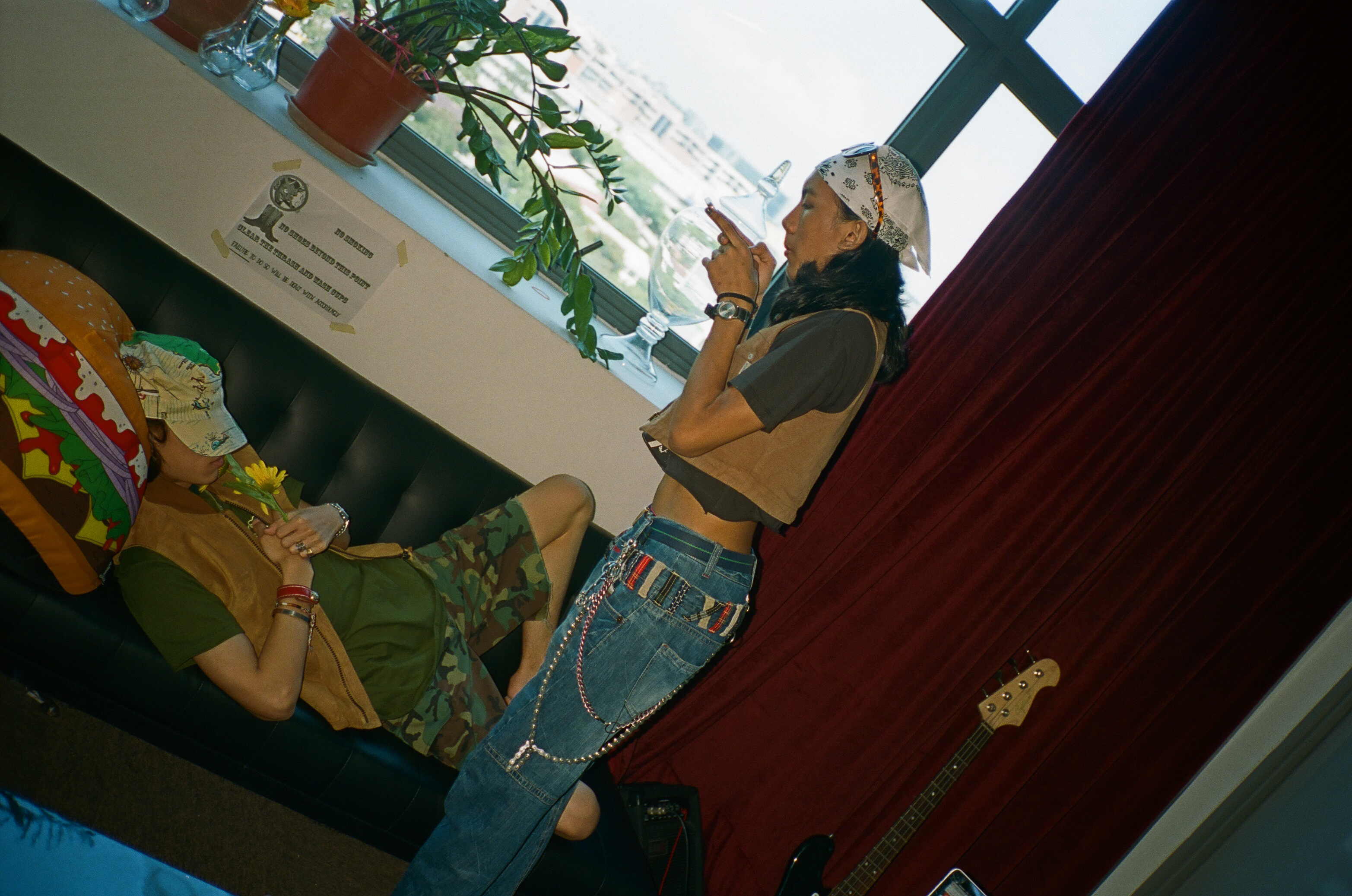 Two people by the studio window, one standing and one reclining beside a flower.