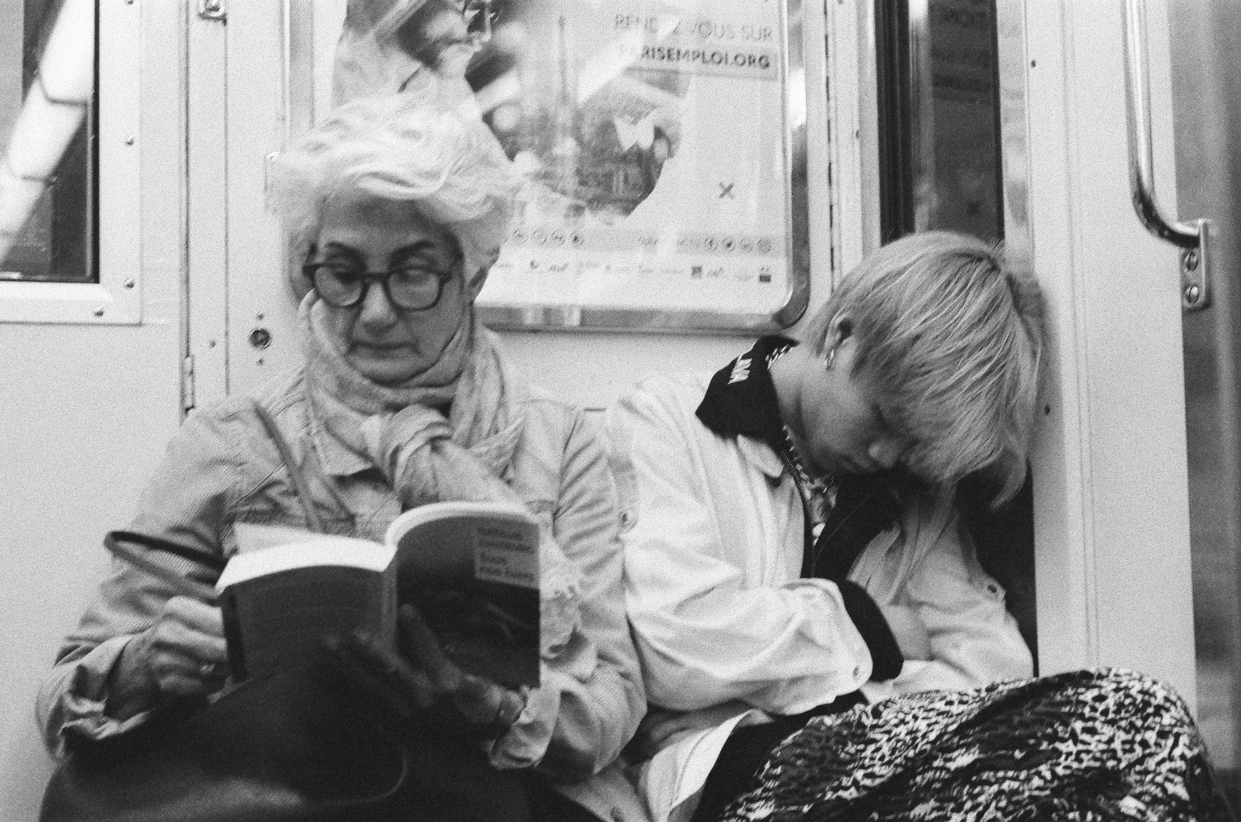 A resting person beside another passenger reading on a train.