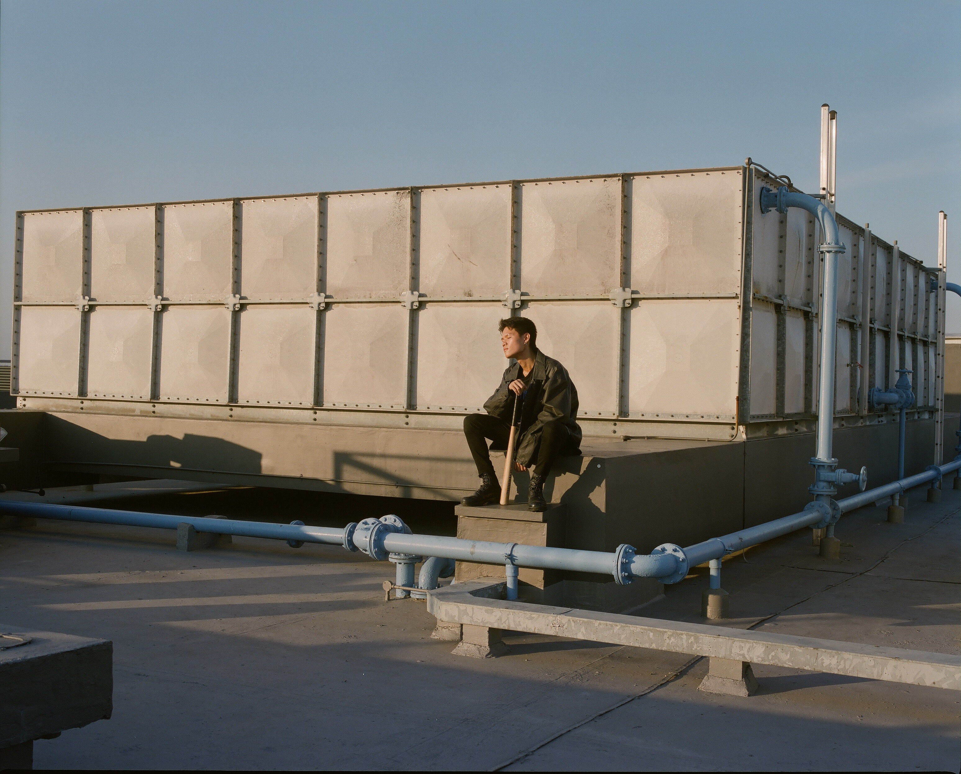 A figure on a rooftop among pipes and walls.