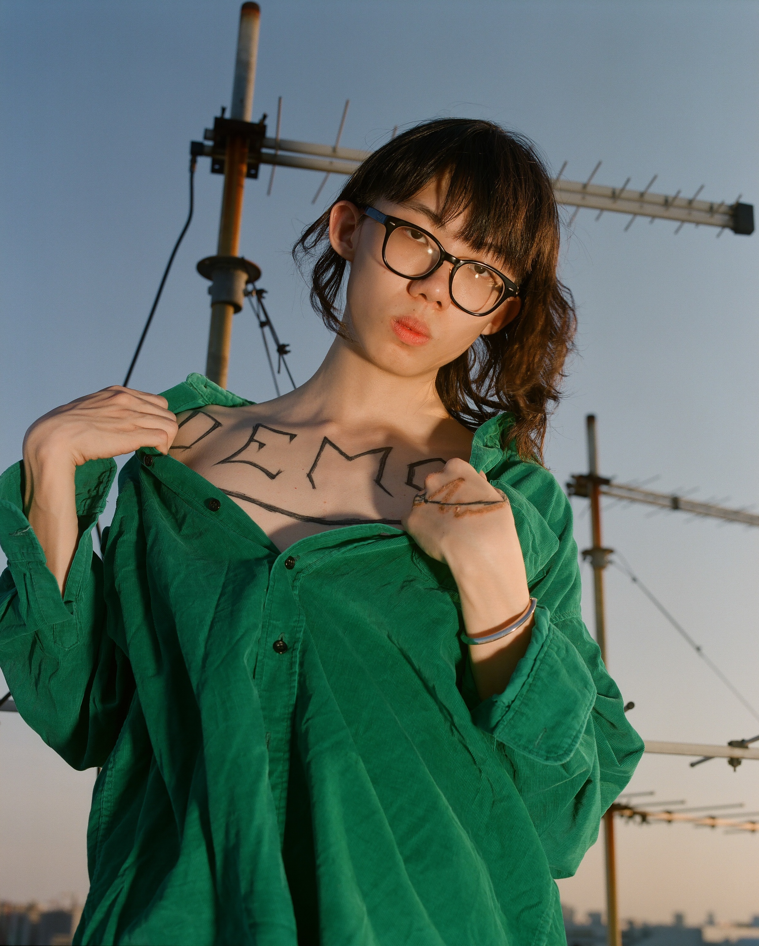 A person in a green shirt and glasses on a rooftop, revealing lettering on their chest.