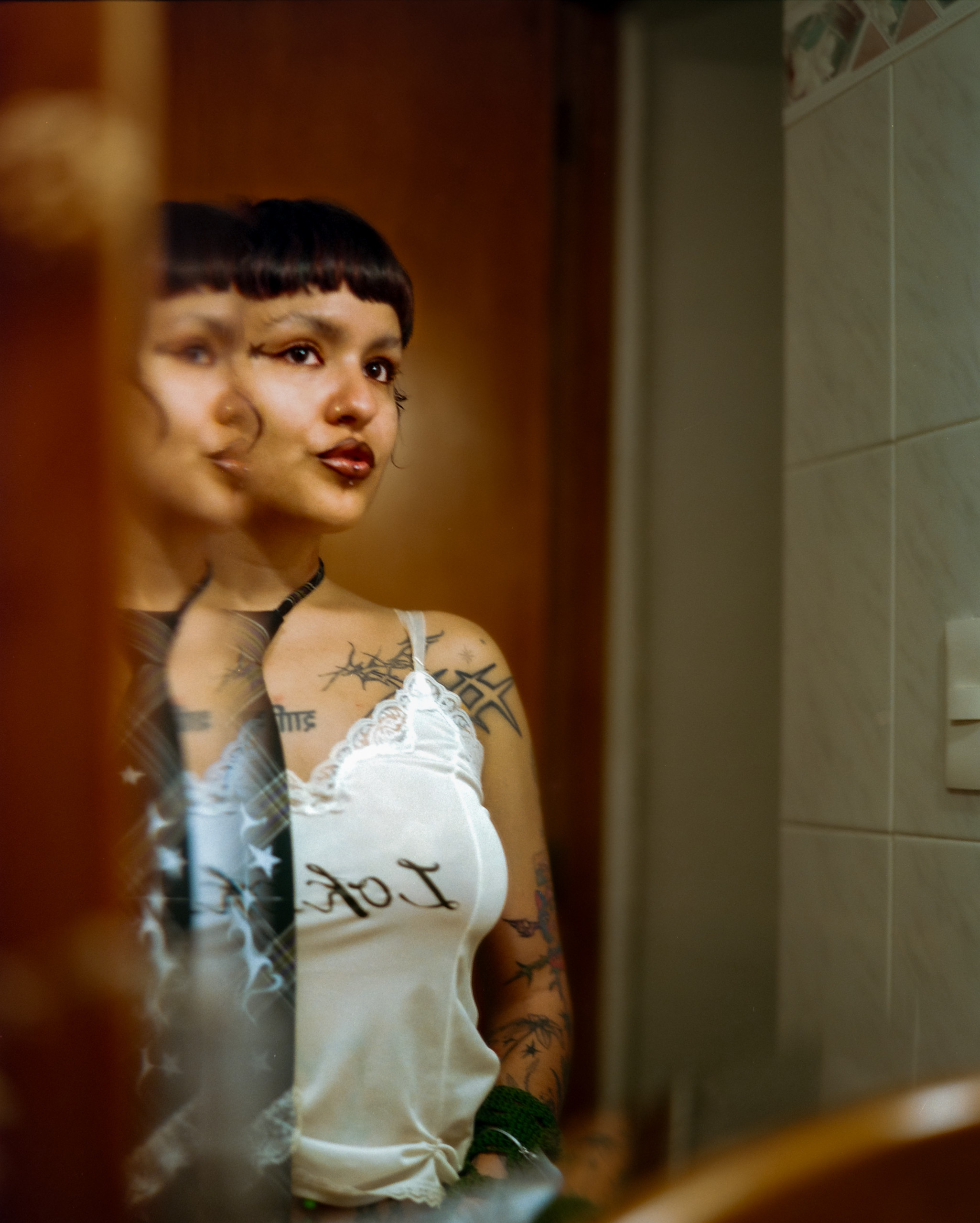 A person with a short fringe seen directly and in reflection inside a tiled room, Mexico City, 2024.