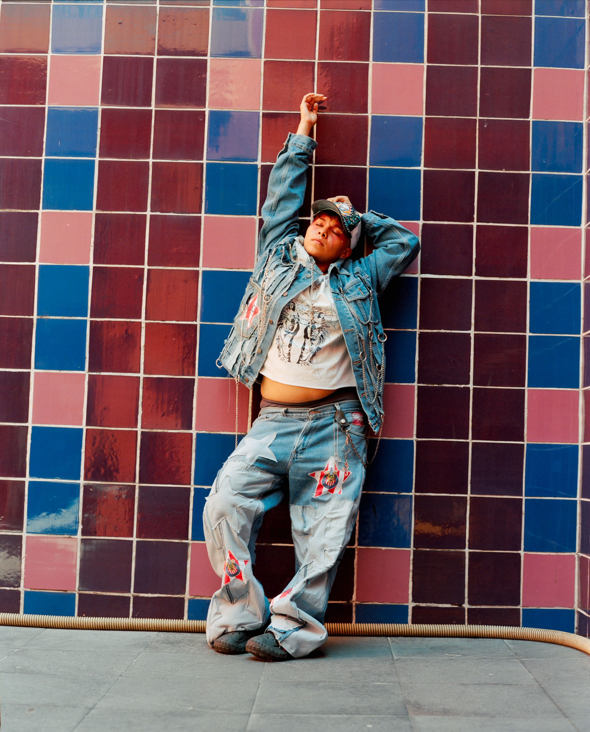 A person raises one arm while standing against a wall of red and blue tiles, Mexico City, 2024.