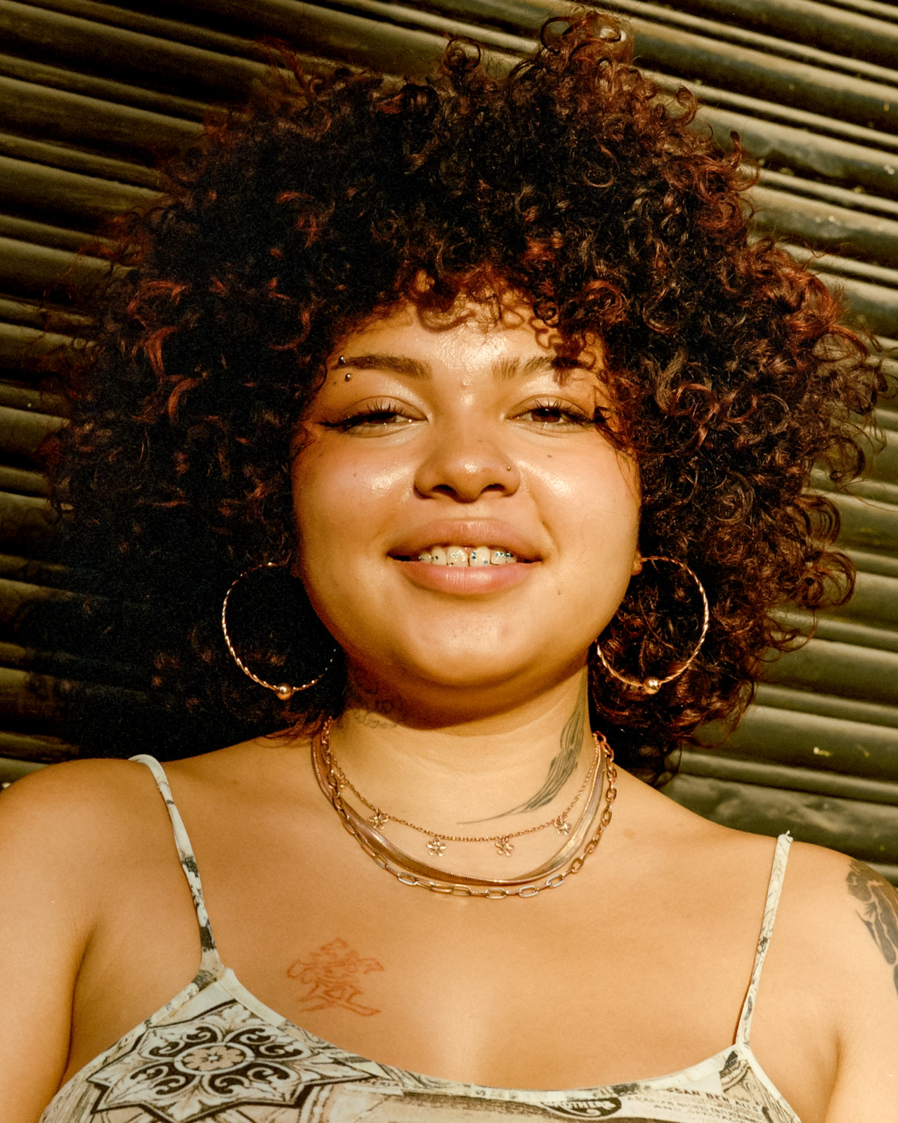 Smiling portrait of a person with curly hair in warm sunlight before a metal shutter, Mexico City, 2024.