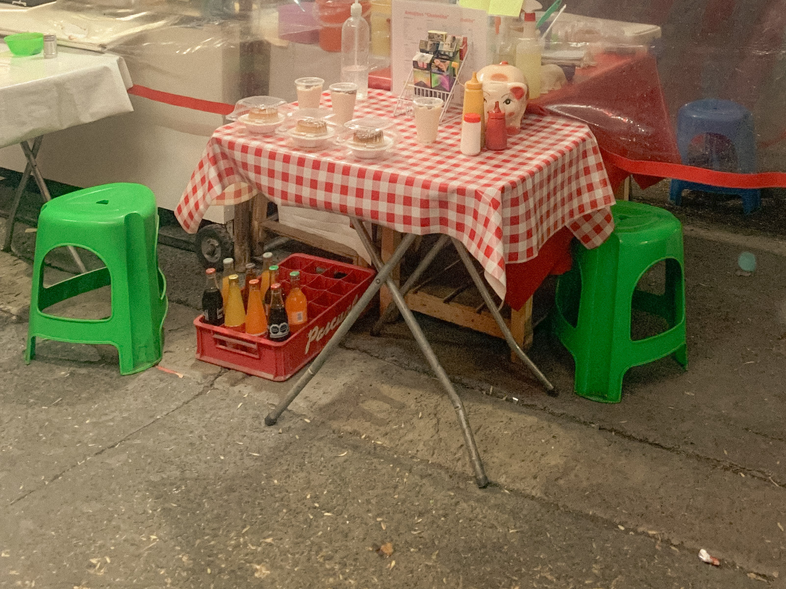 A small street table with a red-and-white checked cloth, bottles and bright green stools, Mexico City, 2024.