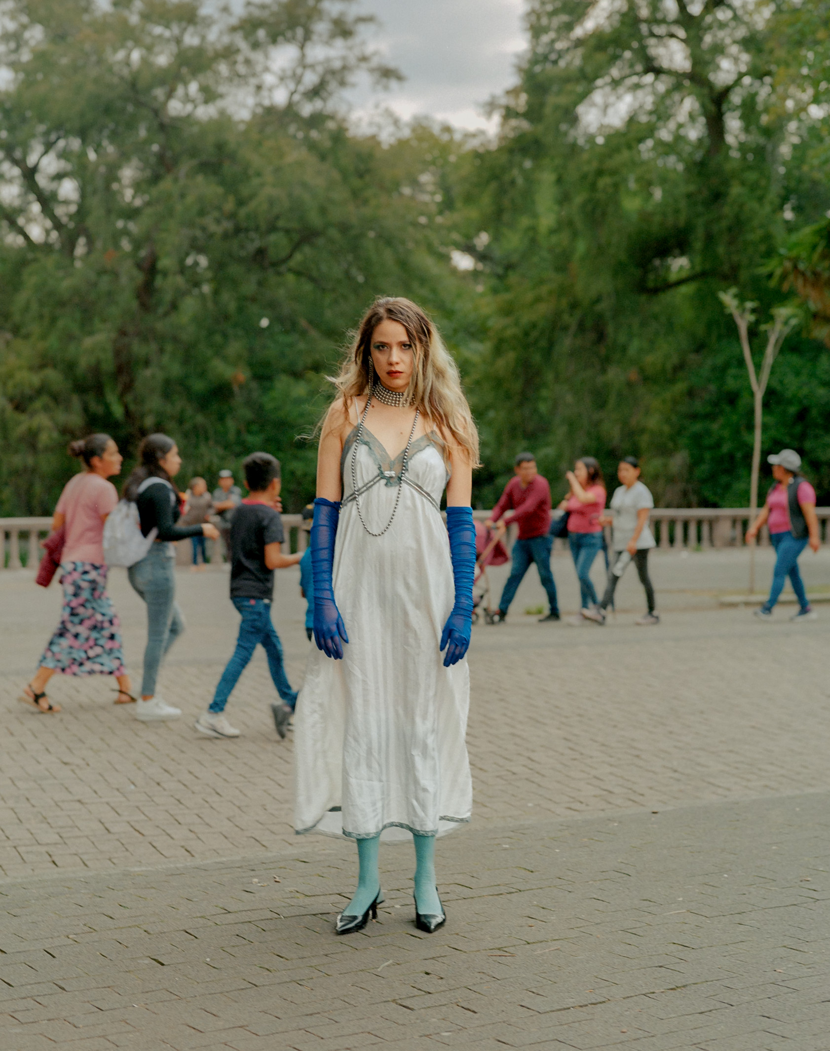 A person in a white dress and blue gloves stands in a public square as others walk behind, Mexico City, 2024.