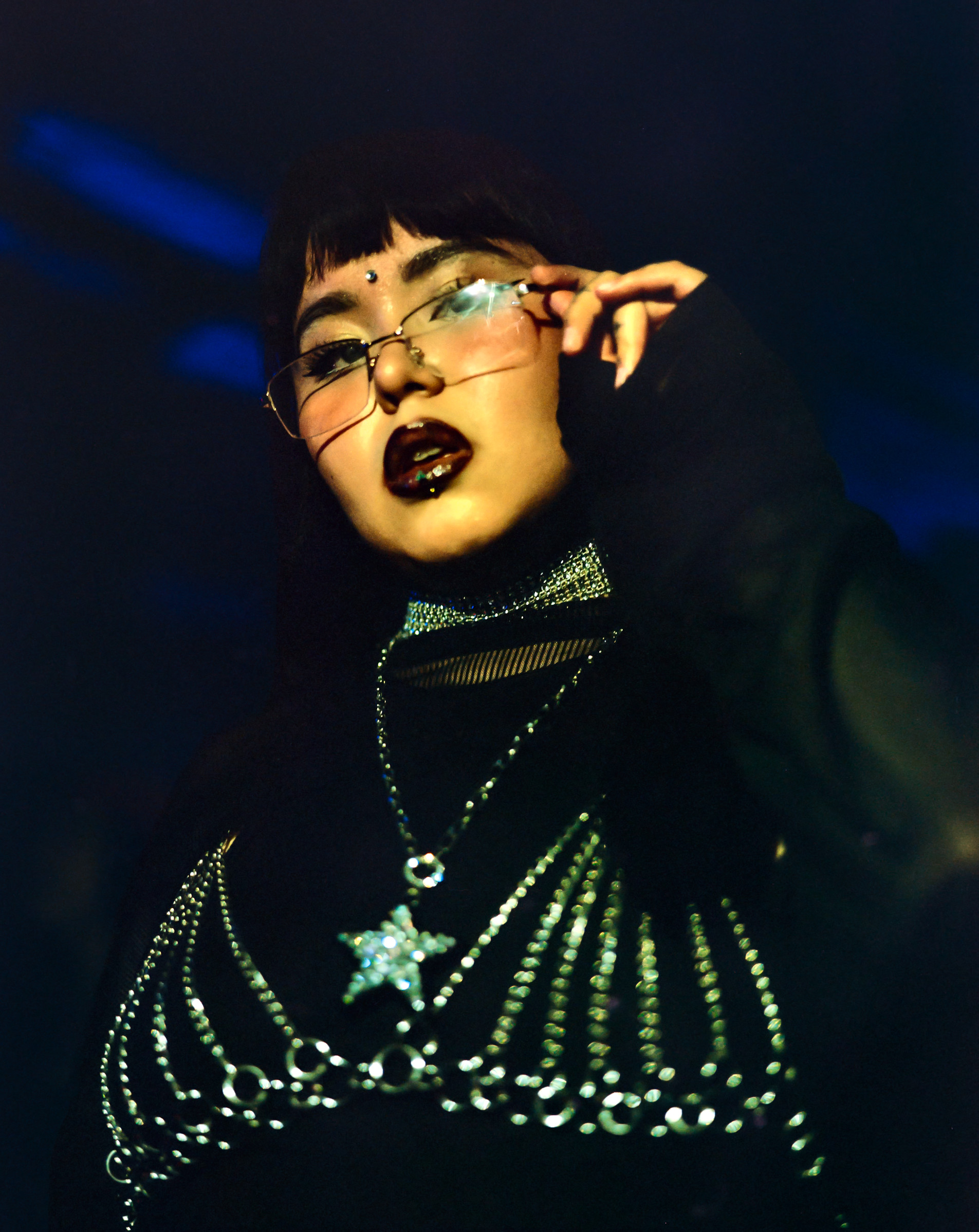 A performer in black reaches towards their glasses under blue stage light, Mexico City, 2024.