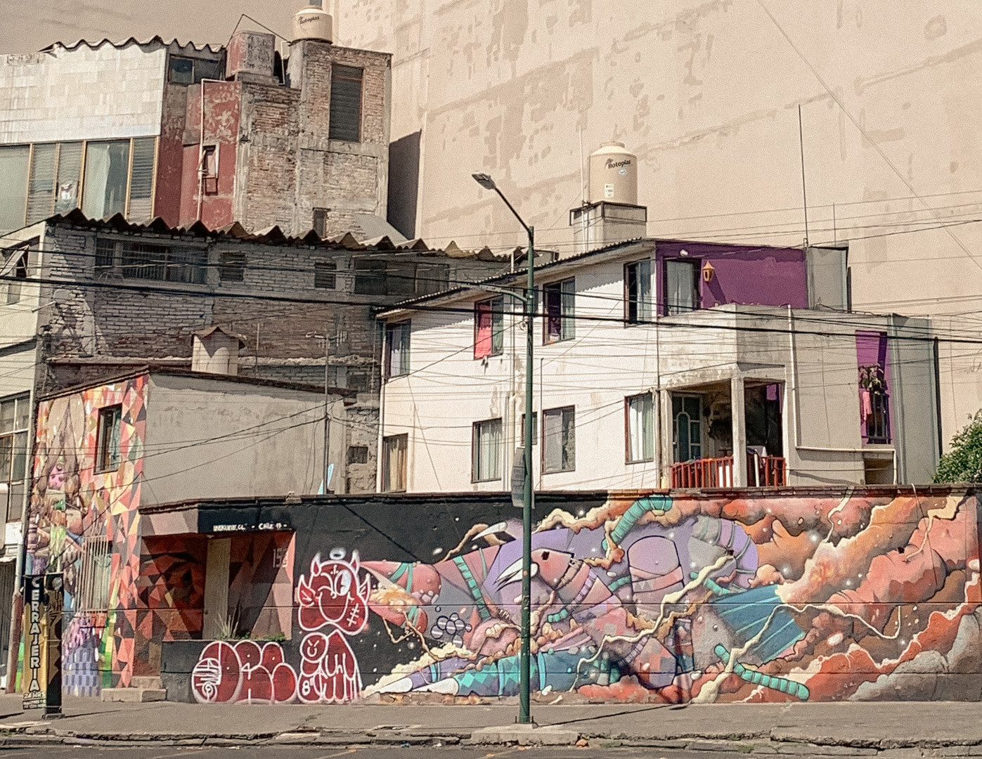 Urban buildings behind a long wall covered with a painted mural and graffiti, Mexico City, 2024.