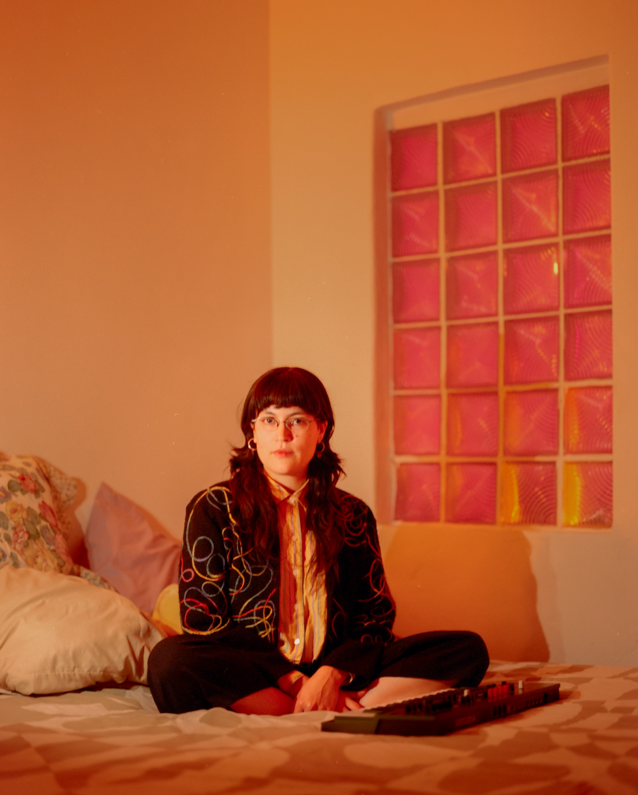 A person seated on a bed in an orange-lit room beside a pink glass-block window, Mexico City, 2024.
