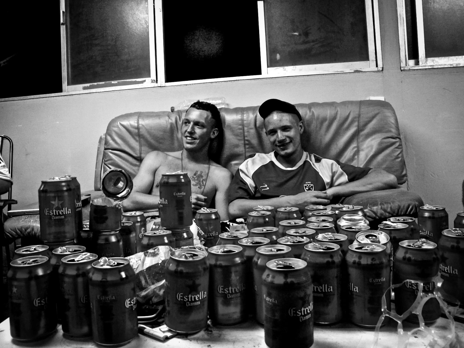 Black-and-white photograph: two people at a table covered with cans and bottles.