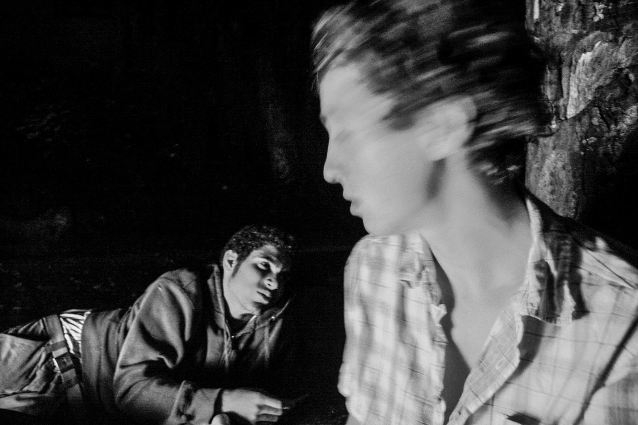 Black-and-white photograph: two people in a dark interior, one moving through the foreground.