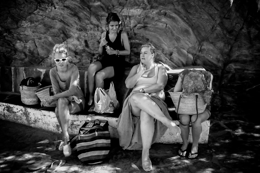 Black-and-white photograph: three people sitting outside against a stone wall.