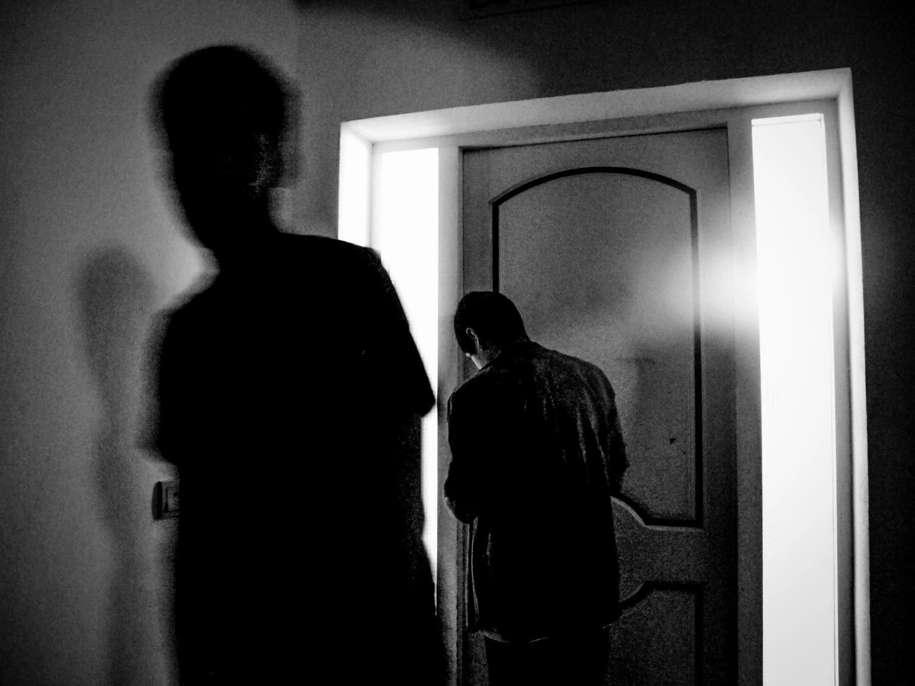 Black-and-white photograph: a silhouette beside a doorway, with another person beyond.