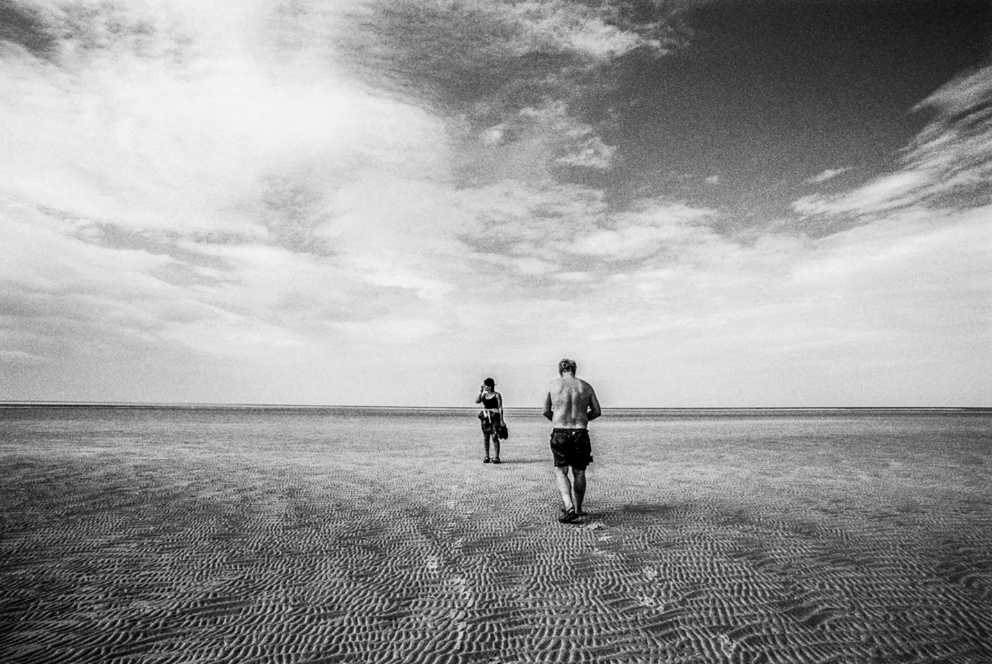 Black-and-white photograph: two people walking across a wide beach.