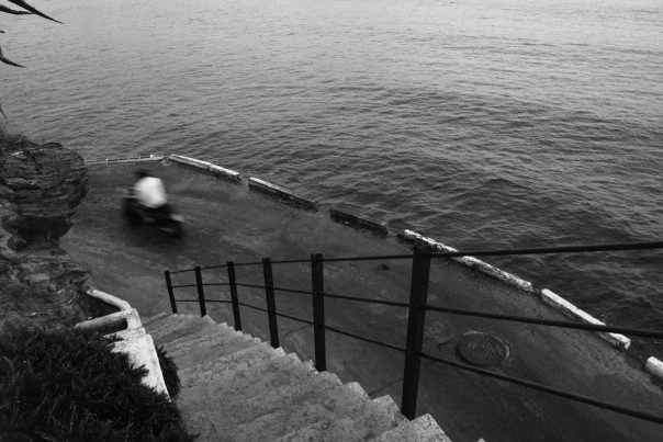 Black-and-white photograph: a person bathing beside steps leading into the sea.