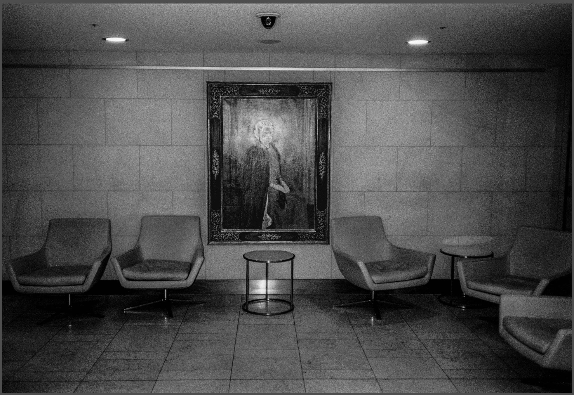 Black-and-white photograph: empty seats in a waiting room beneath a framed portrait.