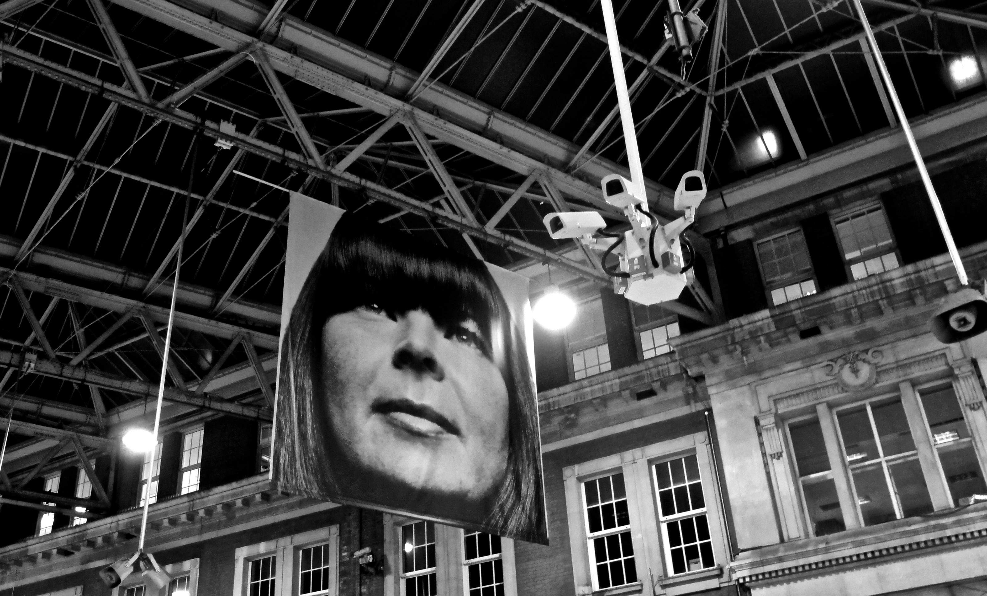 Black-and-white photograph: a large portrait hanging beneath a station roof.