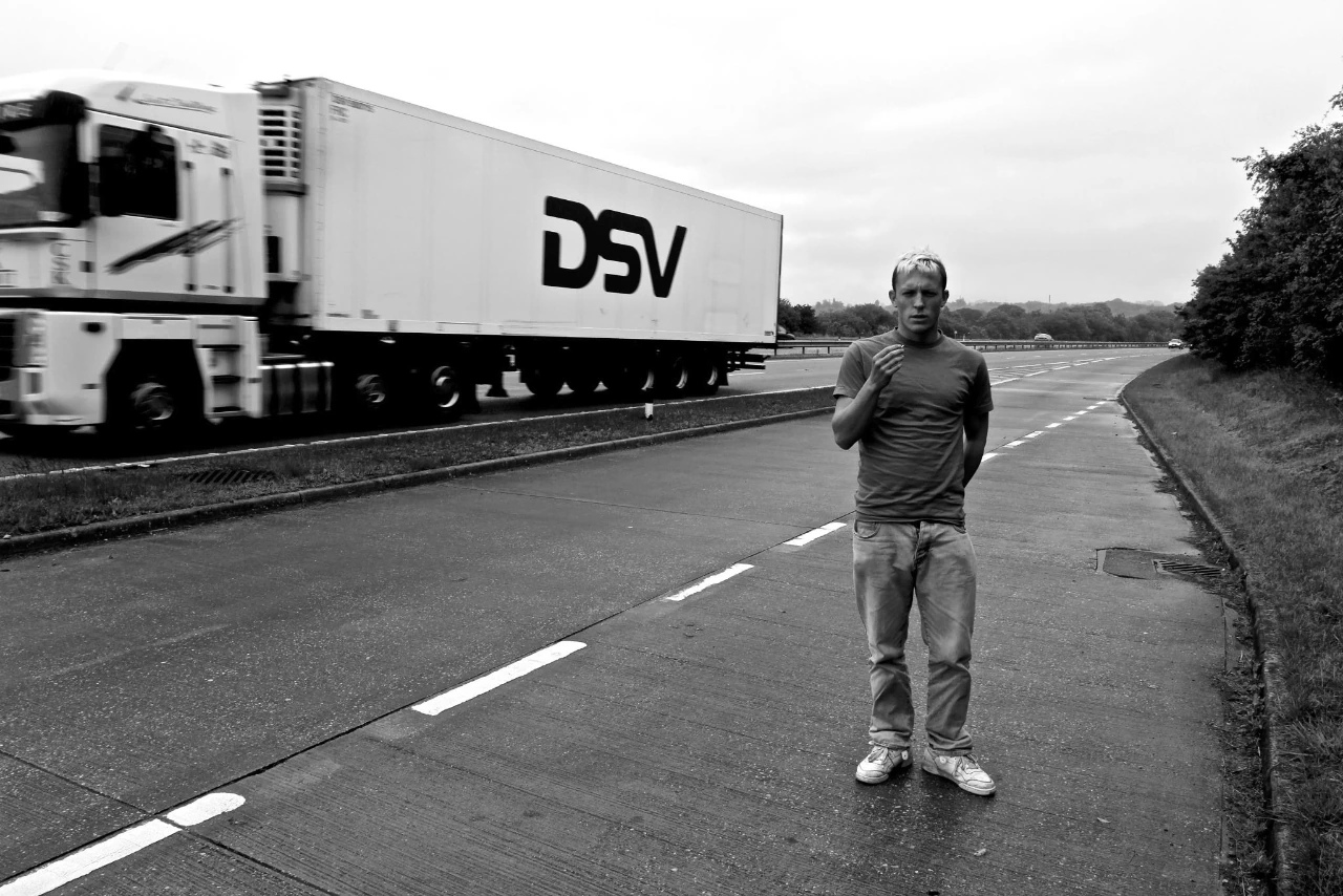 Black-and-white photograph: a person standing beside a road as a lorry passes.