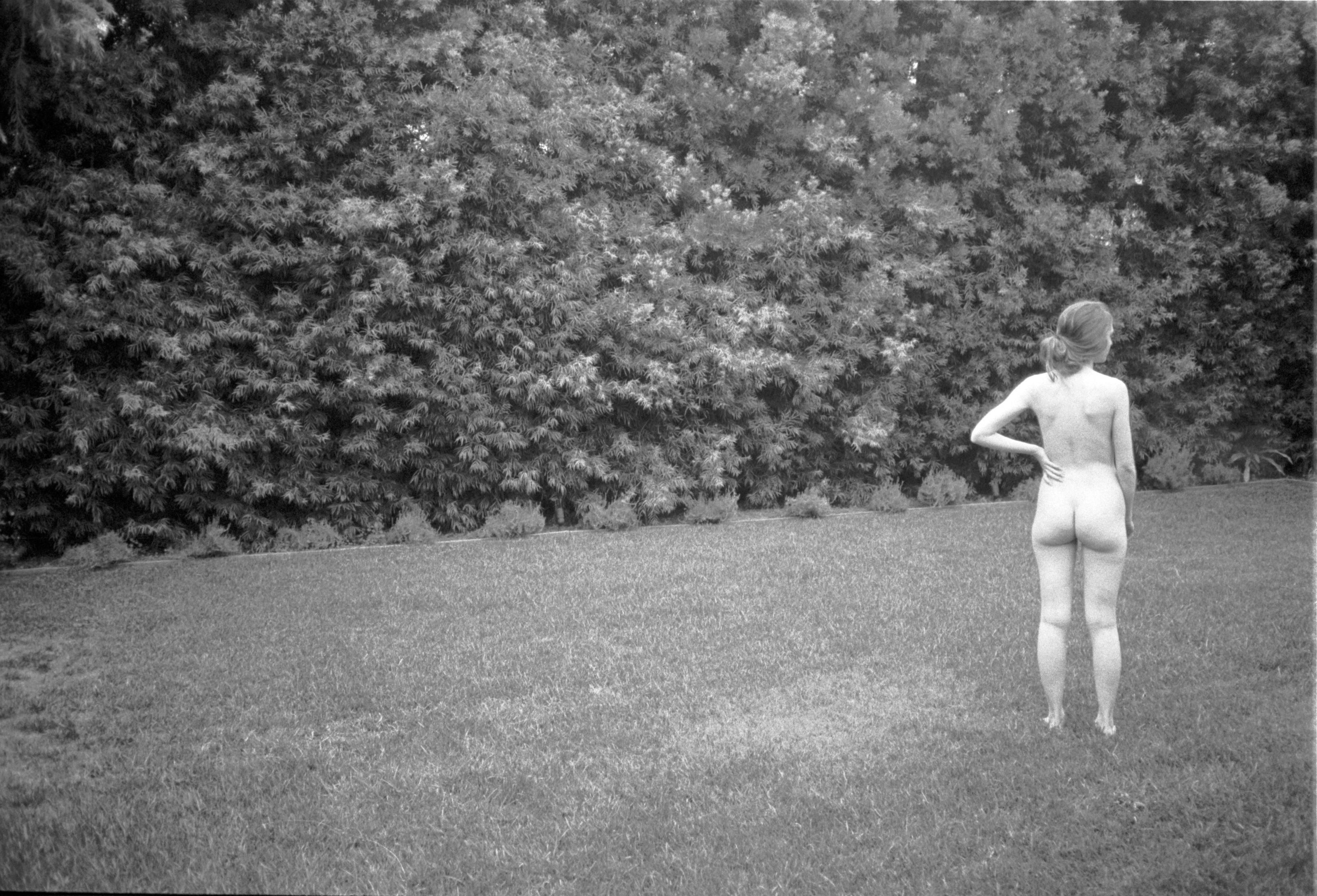 Black-and-white photograph: a nude figure seen from behind at the edge of a garden.