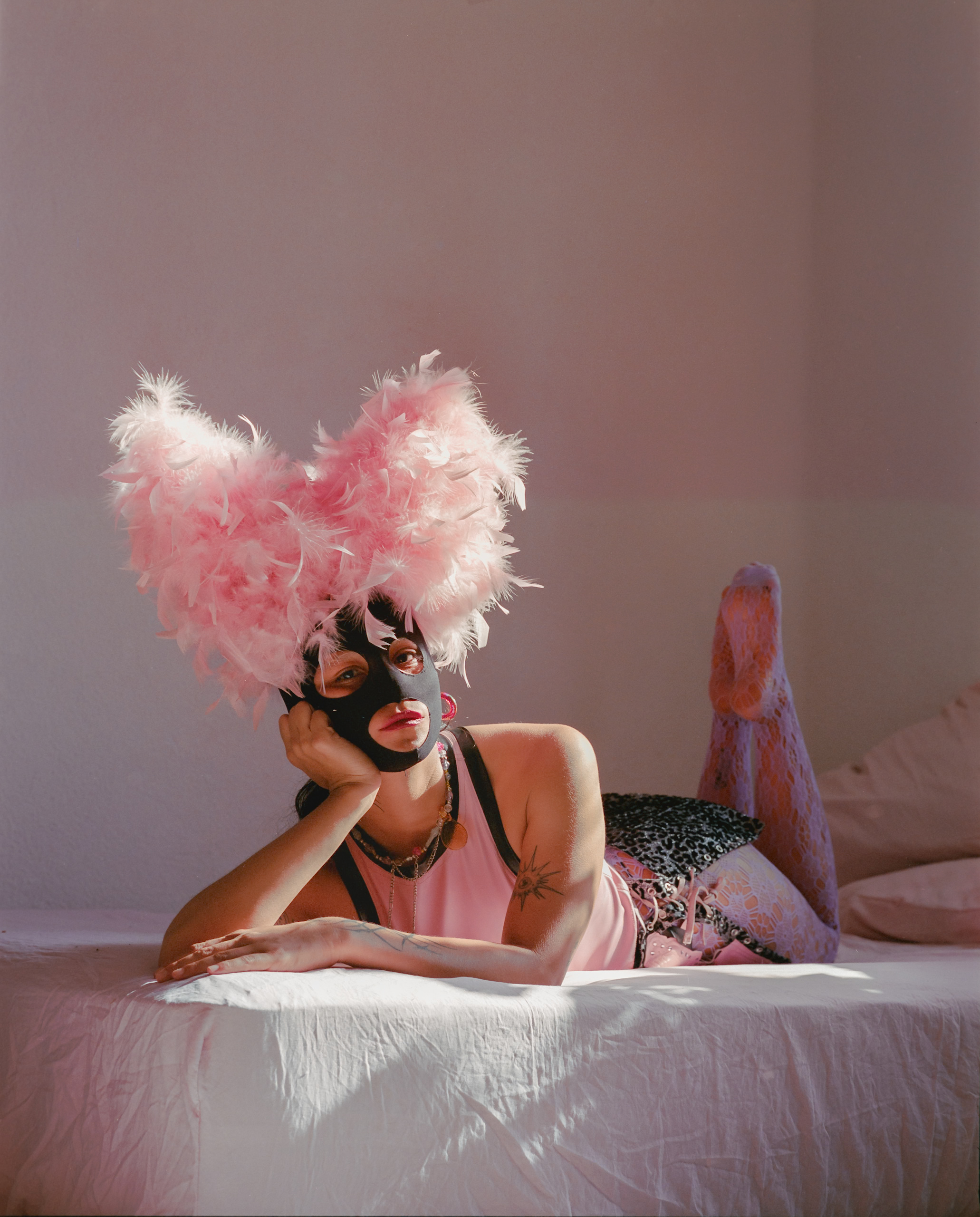 A reclining figure wearing a pink feather headdress on a bed.