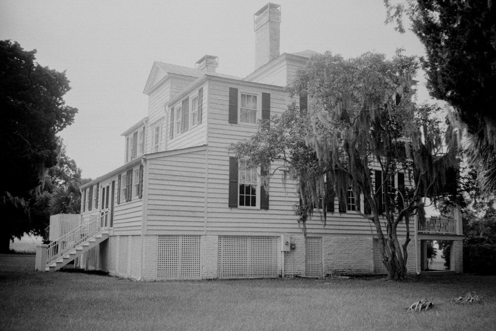 A black-and-white photograph of a wooden house behind a fence.
